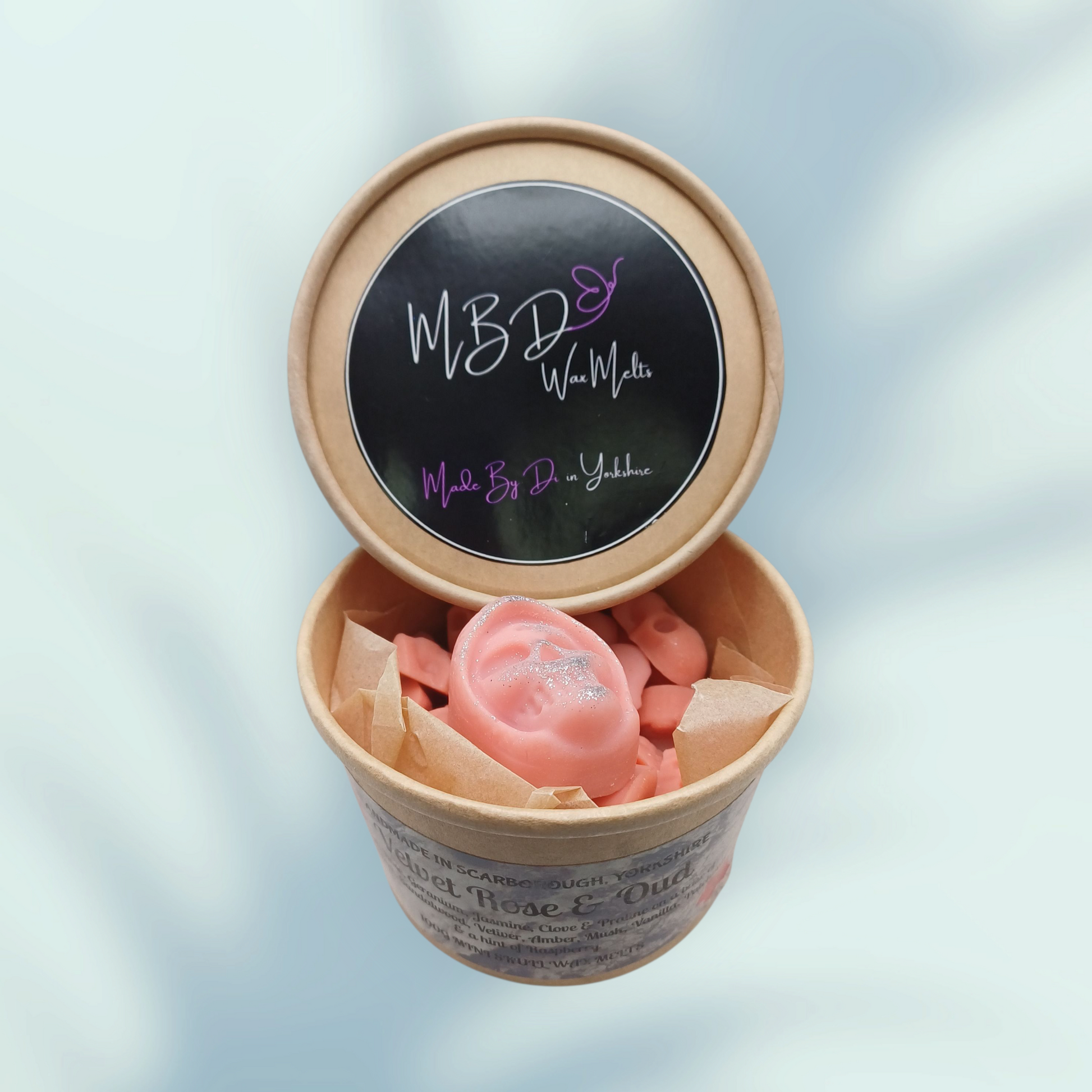 Small cardboard container with pink mini wax melts and a visible brand logo on a light blue backgrounds